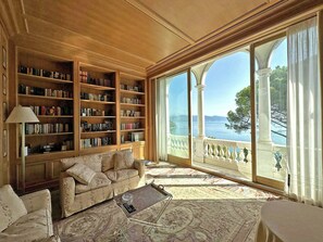 A spacious living room with a large bookshelf, a comfortable sofa, a coffee table, and a view of the sea through large windows. - Historic villa in Portovenere on the Gulf of Poets (Portovenere)
