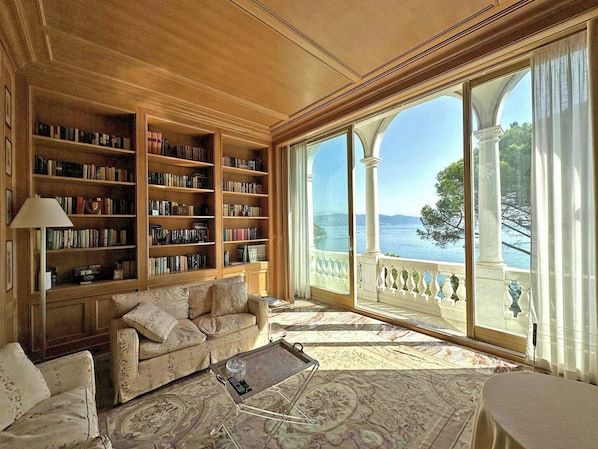 Ein geräumiges Wohnzimmer mit einer großen Bücherwand, einem bequemen Sofa, einem Couchtisch und Meerblick durch große Fenster.