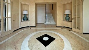 Um corredor com piso de mármore, um design geométrico central em preto e branco, e uma escadaria que leva a outra área.