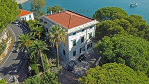 Um edifício de vários andares com telhado de telhas vermelhas, rodeado por vegetação exuberante e vista para o mar.