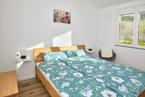 Een slaapkamer met een houten bed, een nachtkastje, een stoel, een raam met uitzicht op bomen en een wandlamp.