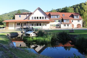 Ein großes, zweistöckiges Haus mit Teich davor, umgeben von Grünflächen und Hügeln.