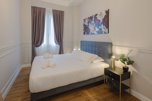 Ein ordentlich bezogenes Bett mit weißen Leinen und gerollten Handtüchern, ein blau gepolsterter Kopfteil, ein Nachttisch mit Lampe und Topfpflanze sowie ein Blumenbild an der Wand.