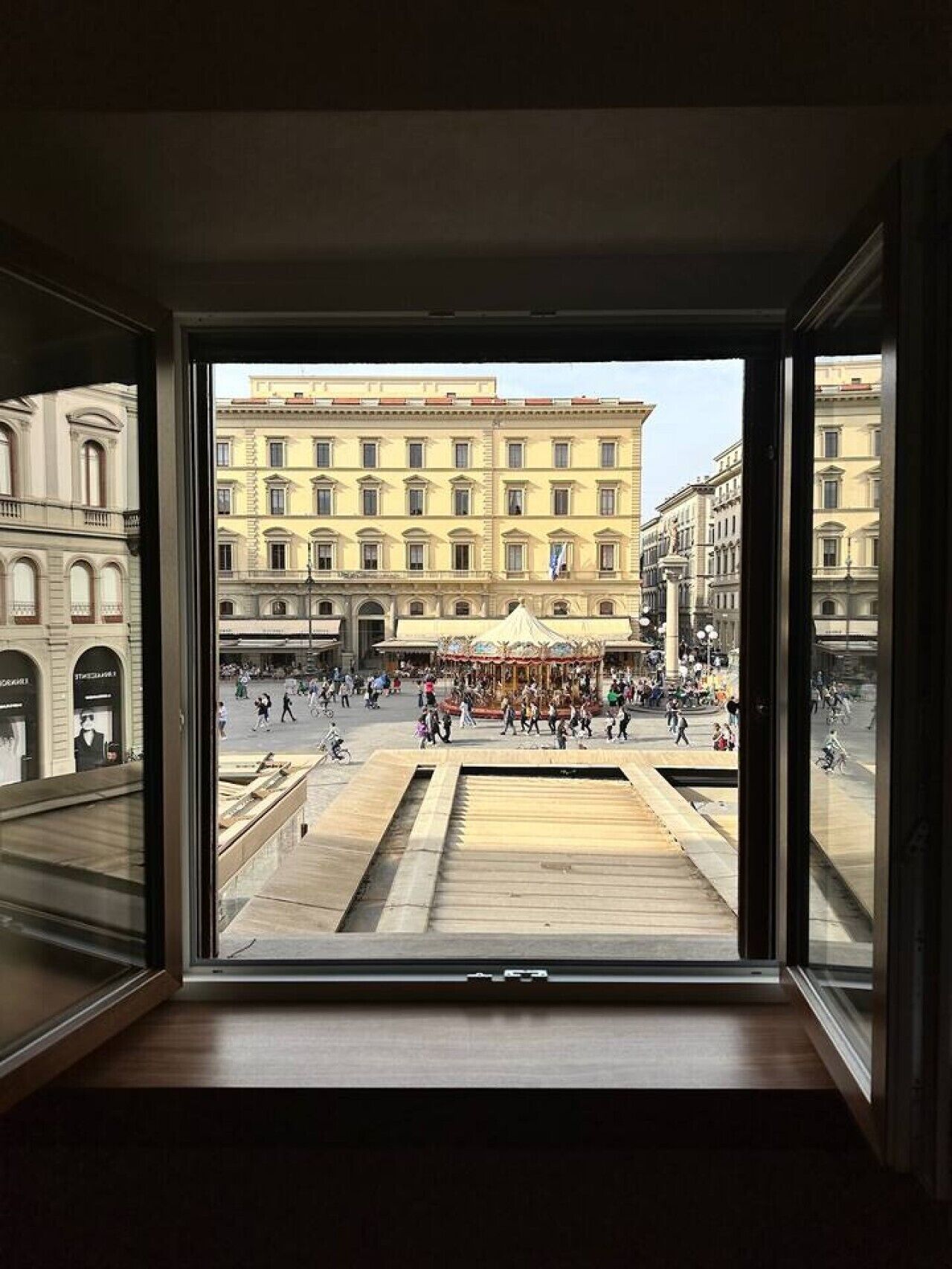 Vista da una stanza con una grande finestra che si affaccia su una piazza con edifici storici, una fontana e una folla.