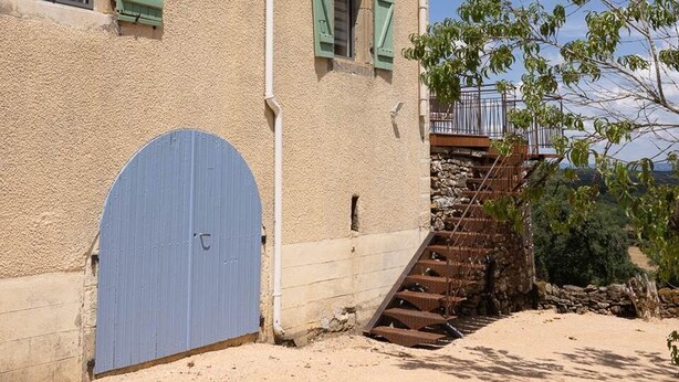 Un edificio de dos pisos con una puerta azul, una escalera y vistas del paisaje circundante.