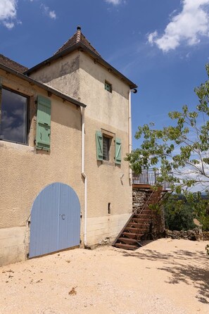 A two-story building with a blue door, a staircase, and a view of the surrounding landscape. - In the Shadow of First Names - Alice's Guesthouse (Miers)