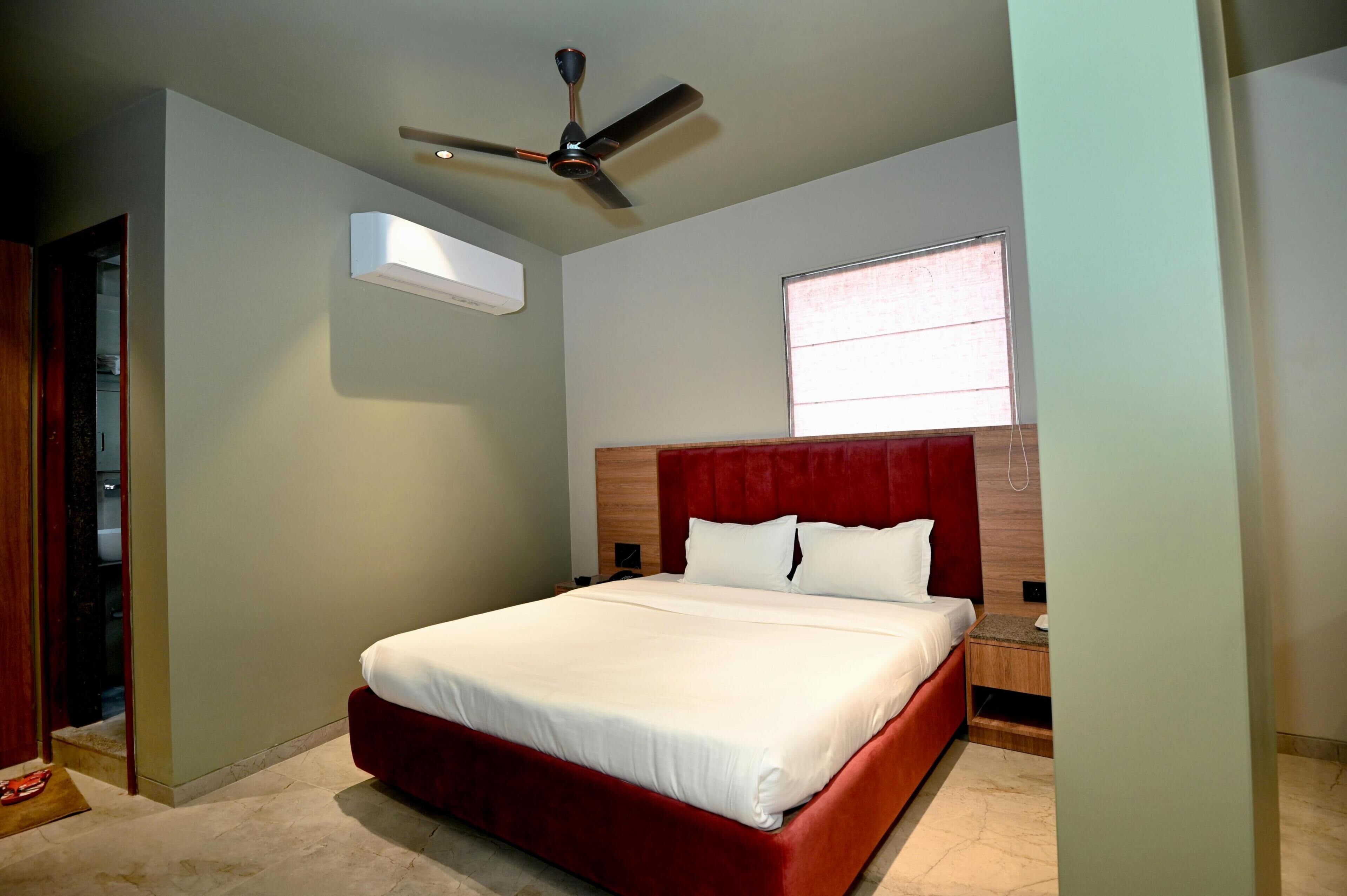A bedroom with a bed, a ceiling fan, an air conditioning unit, a nightstand, and a window.