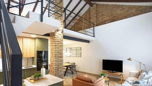 Un loft moderno con cocina, comedor y sala de estar, con ladrillo visto, una escalera y un televisor grande.