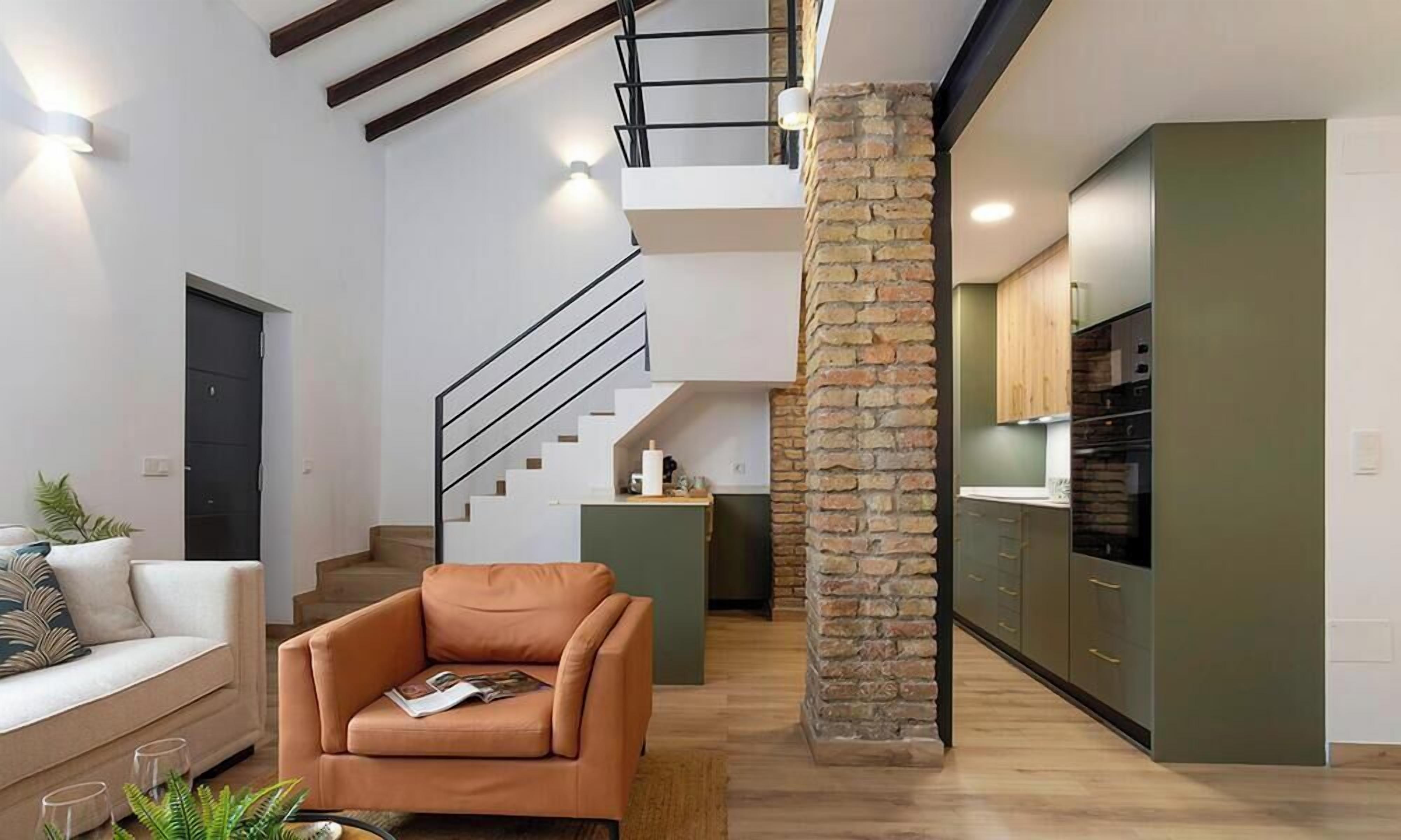 Um interior moderno com uma escada, uma sala de estar com sofá e poltrona, e uma cozinha com armários verdes.