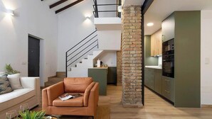 Un interior moderno con una escalera, una sala de estar con sofá y sillón, y una cocina con armarios verdes.