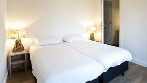 En säng för en person med vita sängkläder, två sänglampor, ett litet träbord och en dörr som leder till ett annat rum.