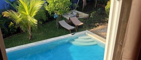 Vista de uma piscina através de uma janela, com uma cerca azul e vegetação ao fundo.