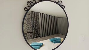 Ein Spiegel mit Zierrahmen, der einen Raum mit einem Bett, Kissen und Wandverkleidung mit vertikalen Lamellen reflektiert.