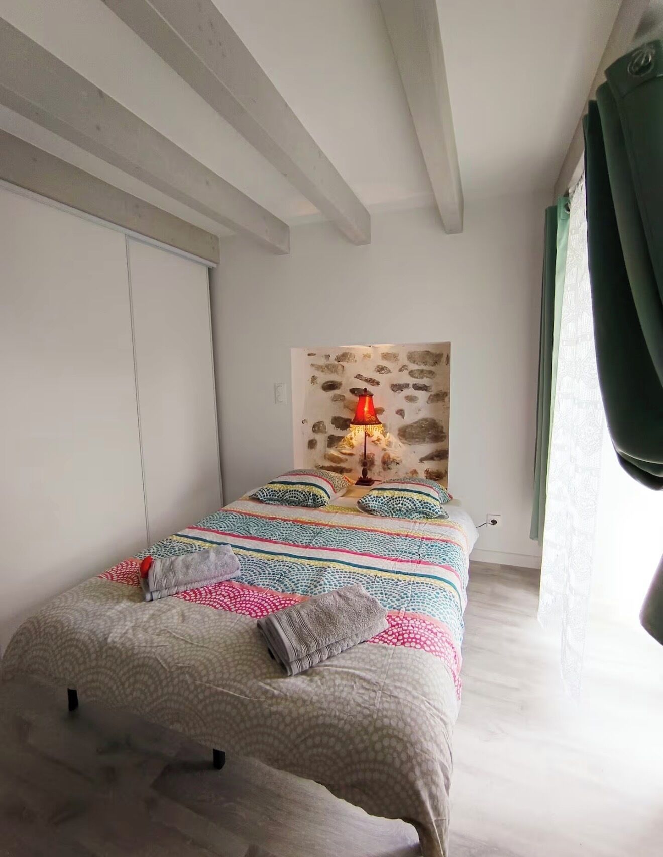 Une chambre à coucher avec un lit, une table de chevet, une lampe, un mur en pierre et une fenêtre avec des rideaux.