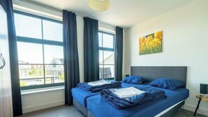 Un dormitorio con cama, cortinas, un cuadro y vistas a edificios.