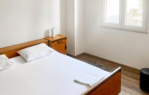 Ein ordentlich bezogenes Bett mit weißen Leinen, ein Nachttisch aus Holz mit einer Lampe und ein Fenster mit Blick ins Grüne.