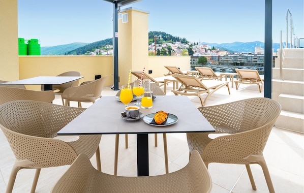 A rooftop terrace with tables and chairs, a croissant, orange juice, and a scenic view of a city and hills. - Awesome apartment in Metkovic with sauna (Metkovic)