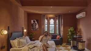A room with a bed, a sofa, a window with curtains, a plant, and a wall-mounted air conditioner. - Minimalist - Lake Pichola Edit (Udaipur)