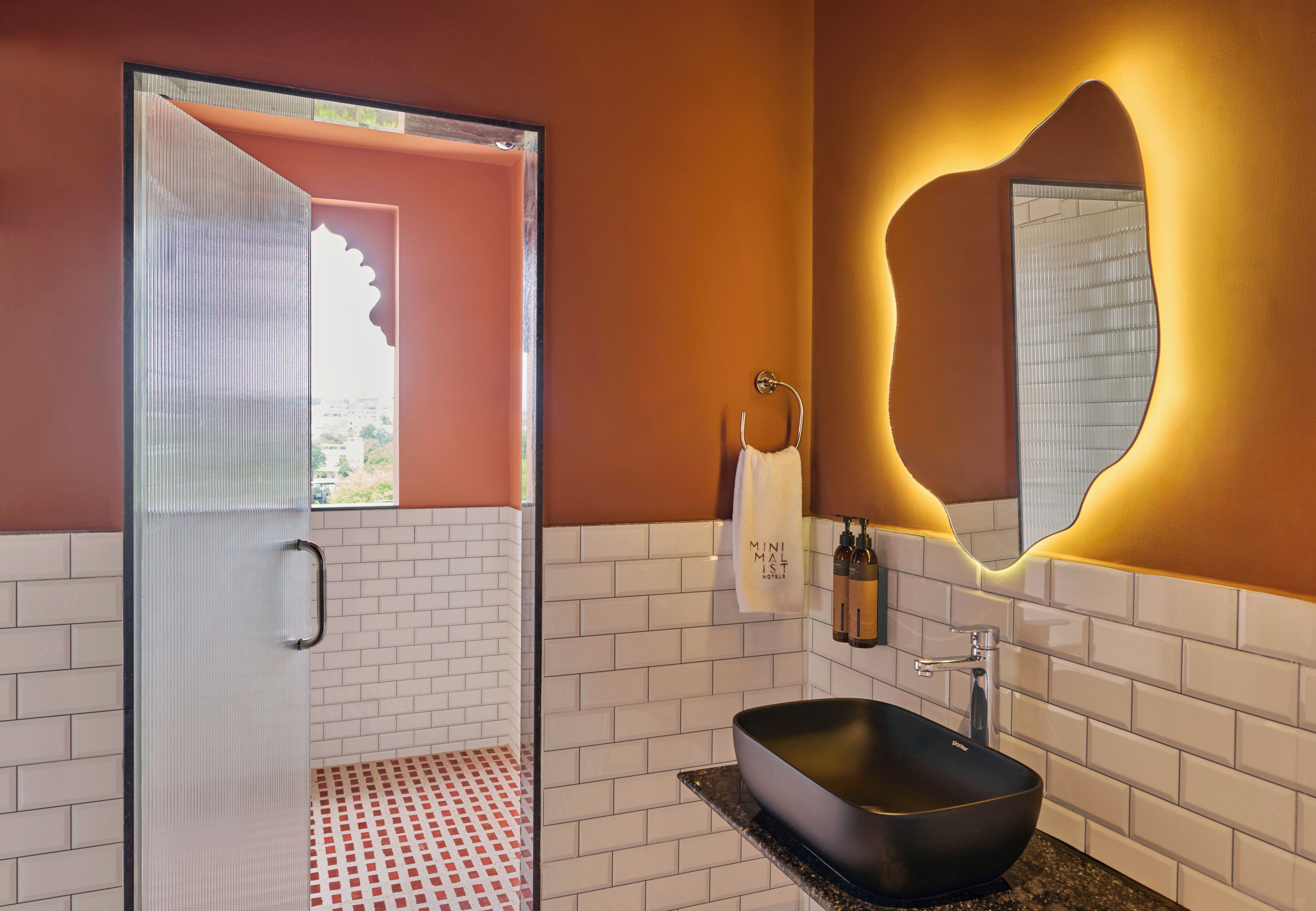 Un baño con mampara de vidrio, lavamanos negro, espejo de forma original y toallero con una toalla blanca.