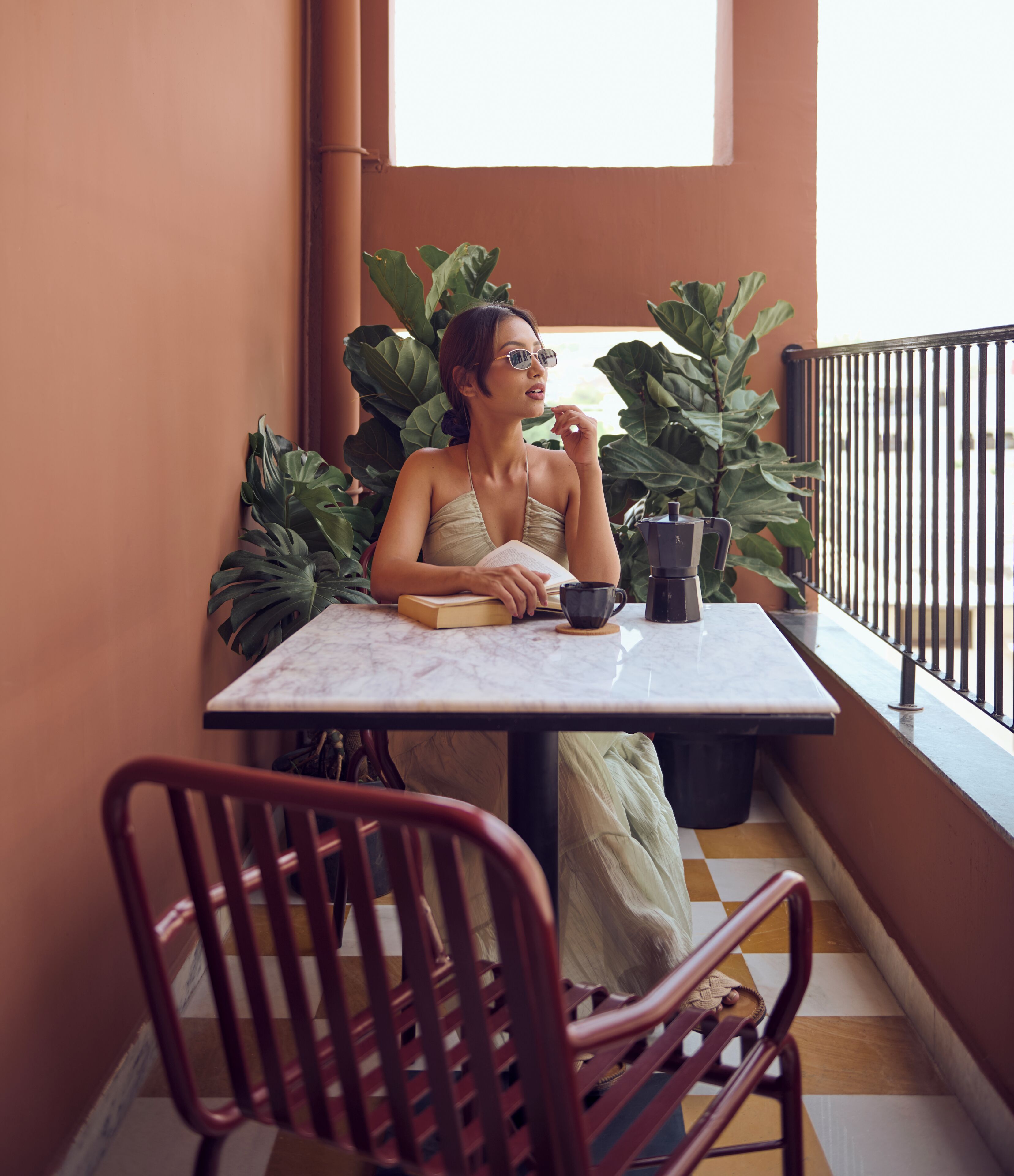 Una persona sentada a una mesa con un libro y una taza, rodeada de grandes plantas verdes, en un balcón con barandilla.