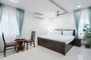 A hotel room with a bed, a desk with chairs, a ceiling fan, an air conditioning unit, and a potted plant.