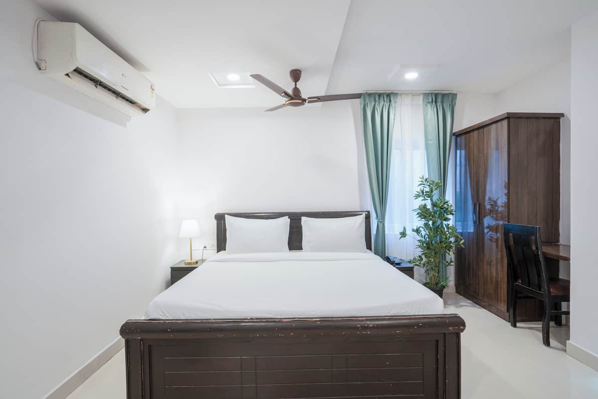 A bedroom with a bed, a ceiling fan, an air conditioning unit, a nightstand with a lamp, a wooden wardrobe, and a potted plant.