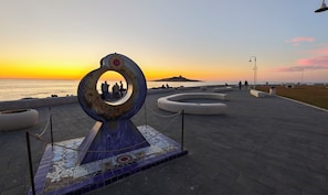 Ein öffentlicher Weg mit einer dekorativen, runden Struktur, Bänken und Blick auf das Meer bei Sonnenuntergang.
