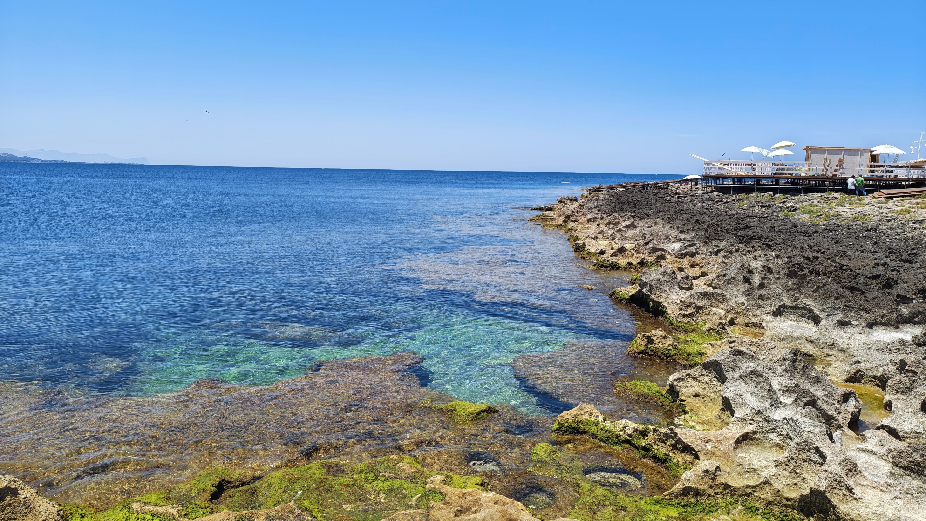 Ein Küstenhotel mit klarem, blauen Wasser, felsigem Gelände und einem Steg mit Sonnenschirmen.