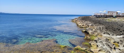 A coastal hotel with clear blue waters, rocky terrain, and a pier with umbrellas.