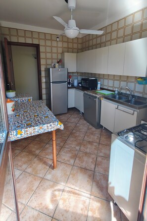Une cuisine avec un sol carrelé, une table recouverte d’une nappe à motifs, un réfrigérateur, un four à micro-ondes, un évier et un ventilateur de plafond.