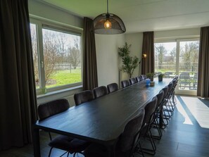 Ein Konferenzraum mit langem Tisch, Stühlen, einer hängenden Leuchte, einem Blumentopf und großen Fenstern mit Vorhängen.