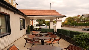 Eine Terrasse im Freien mit Tischen und Stühlen, ein überdachter Sitzbereich und ein Blick auf die Straße und die Gebäude im Hintergrund.