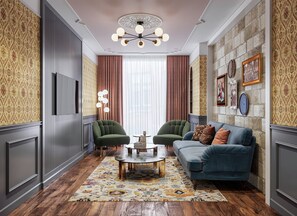 Ein Zimmer mit ornamental gestalteter Tapete, einem grünen Sessel, einem blauen Sofa, einem runden Couchtisch und einem Teppich mit Muster.