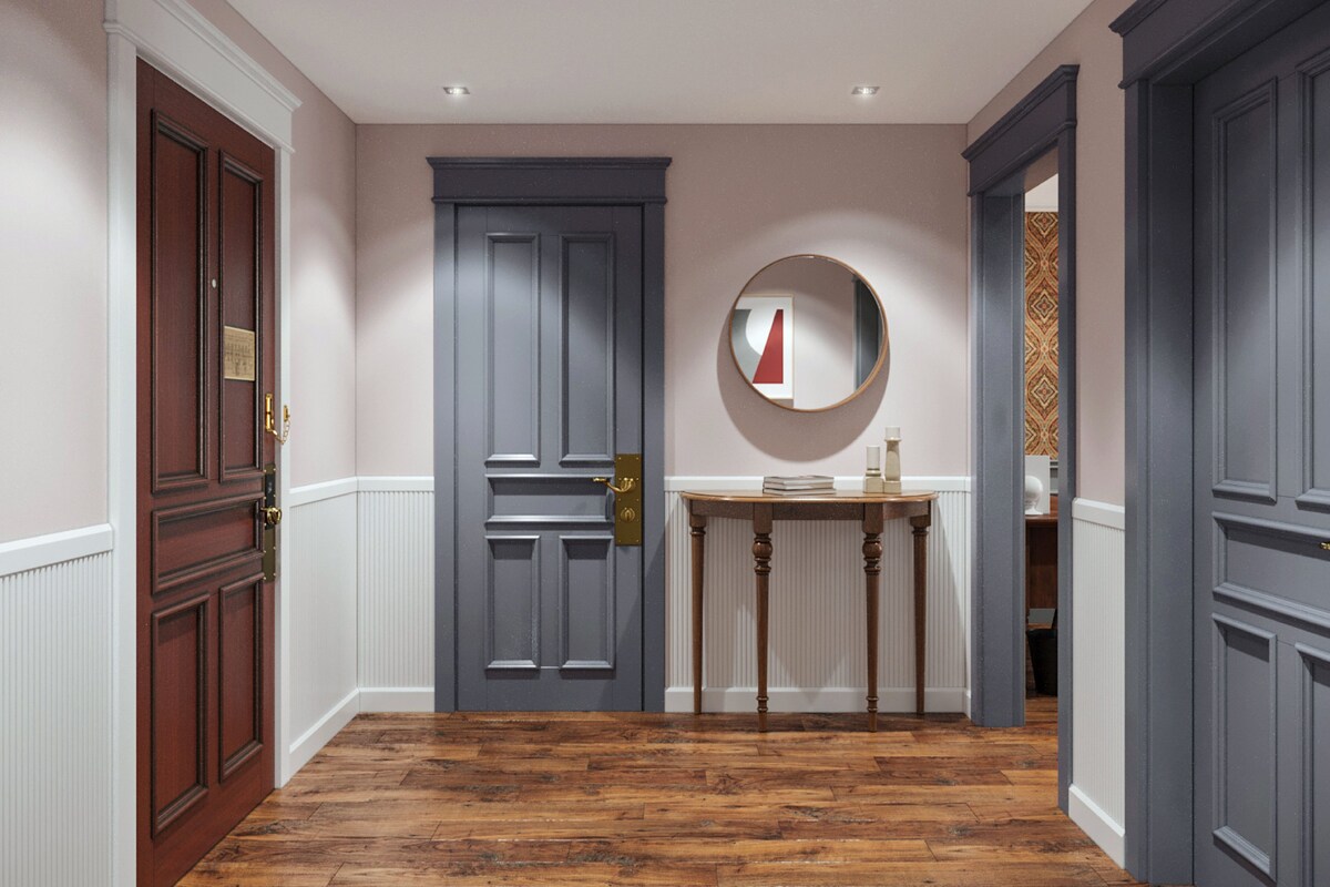 Un couloir avec un plancher en bois, une porte grise, un miroir rond et une petite table sur laquelle sont posés des livres.