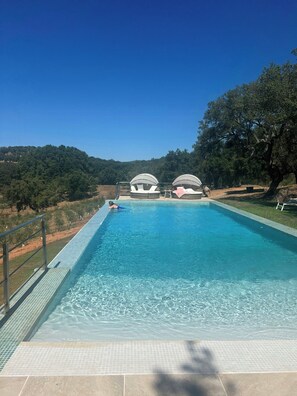 Una piscina rectangular con agua azul cristalina, rodeada por un solárium con baldosas. Al fondo, se observan dos grandes estructuras en forma de cúpula, posiblemente para dar sombra o refugio.