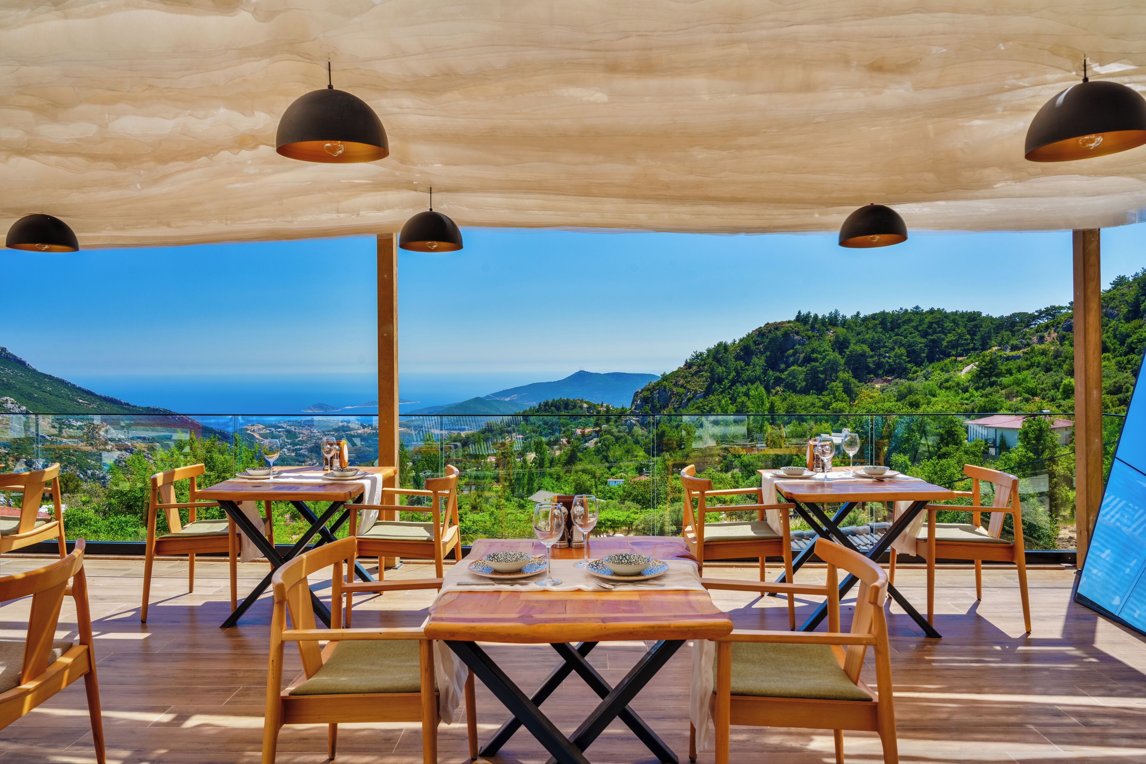 A terrace with wooden tables and chairs, a view of a mountainous landscape, and hanging pendant lights.