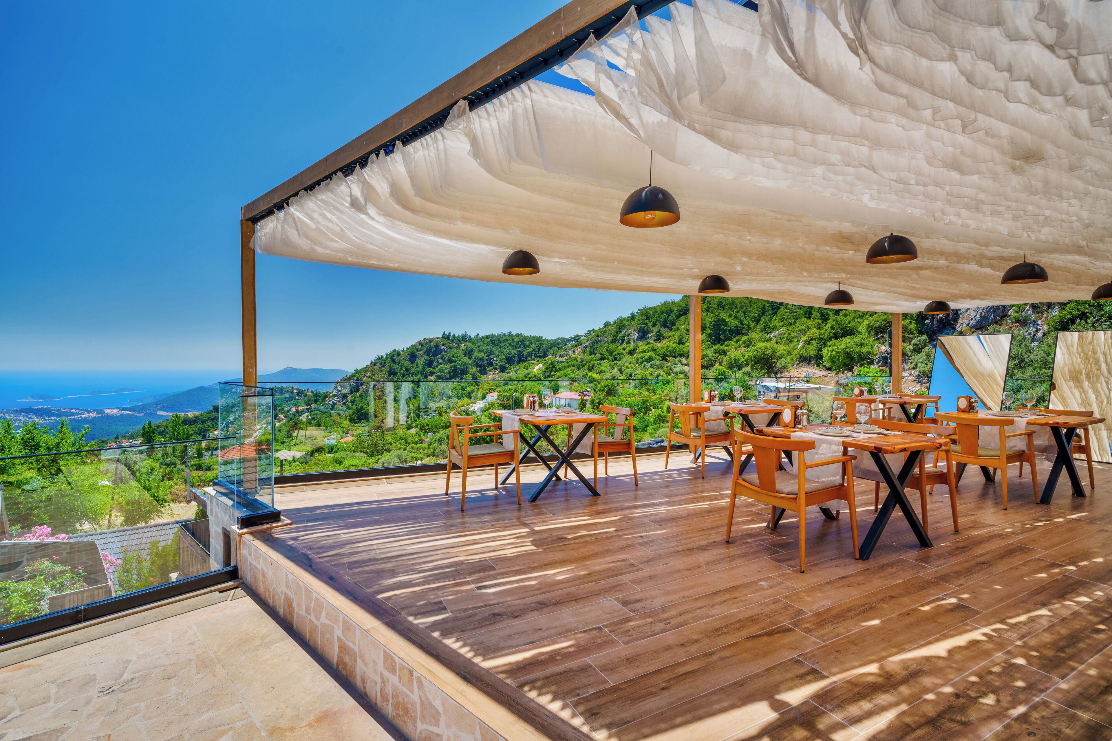 An outdoor terrace with wooden tables and chairs, a white canopy, and a scenic view of a green landscape and mountains.