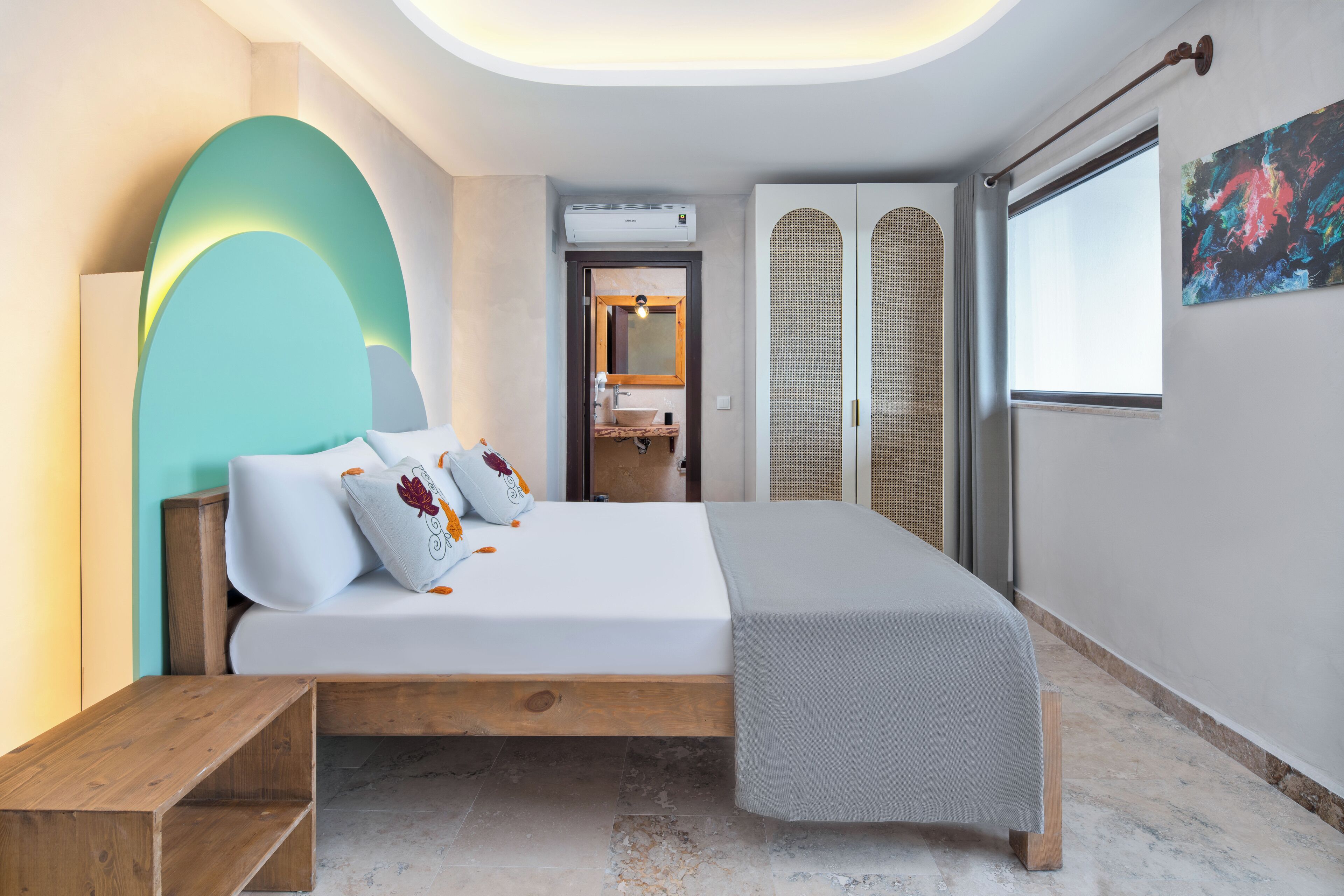 A modern bedroom with a bed, a wooden bench, a bathroom with a mirror, and a window with a view.