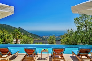 Uma área à beira da piscina com espreguiçadeiras, uma mesa com frutas e bebidas, e vista para o mar e as colinas.