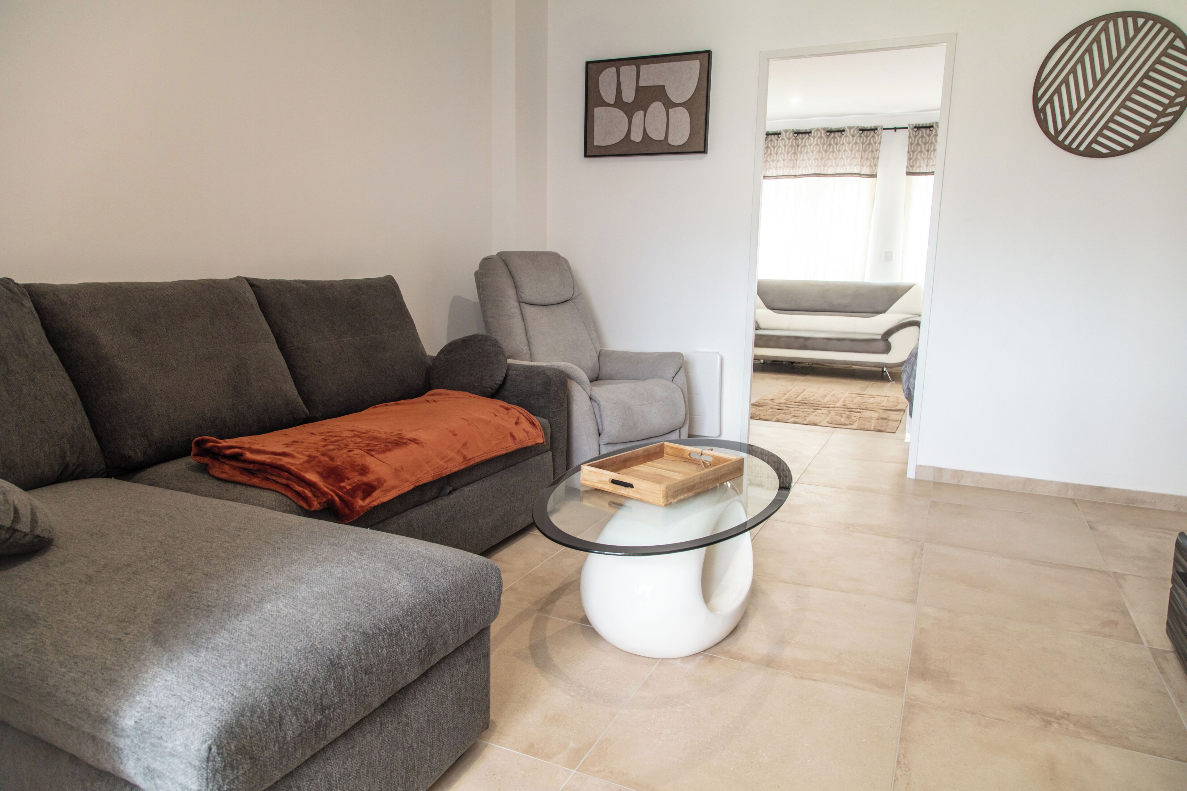 A living room with a grey sofa, a glass coffee table, and a view into another room.