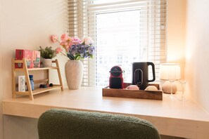 Una habitación con un escritorio de madera, una planta en maceta, un florero con flores, una cafetera y una lámpara.