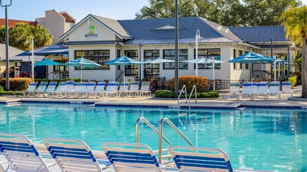 Uma área de piscina com espreguiçadeiras e guarda-sóis, um edifício com uma placa que diz 'Breezes' e um céu azul límpido.