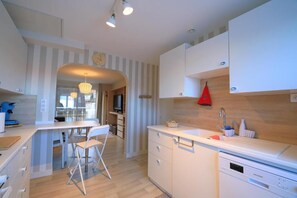 A modern kitchen with white cabinetry, a dining area with a white table and chairs, and a red hanging light. - Les clés de la mer (Le Portel)
