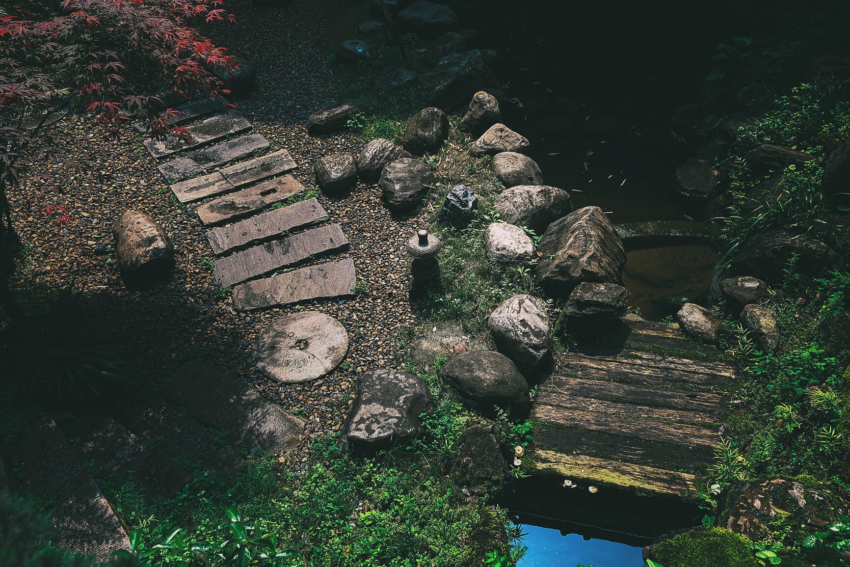 Un sendero de piedra con escalones que conducen a un puente de madera sobre un cuerpo de agua, rodeado de rocas y follaje.