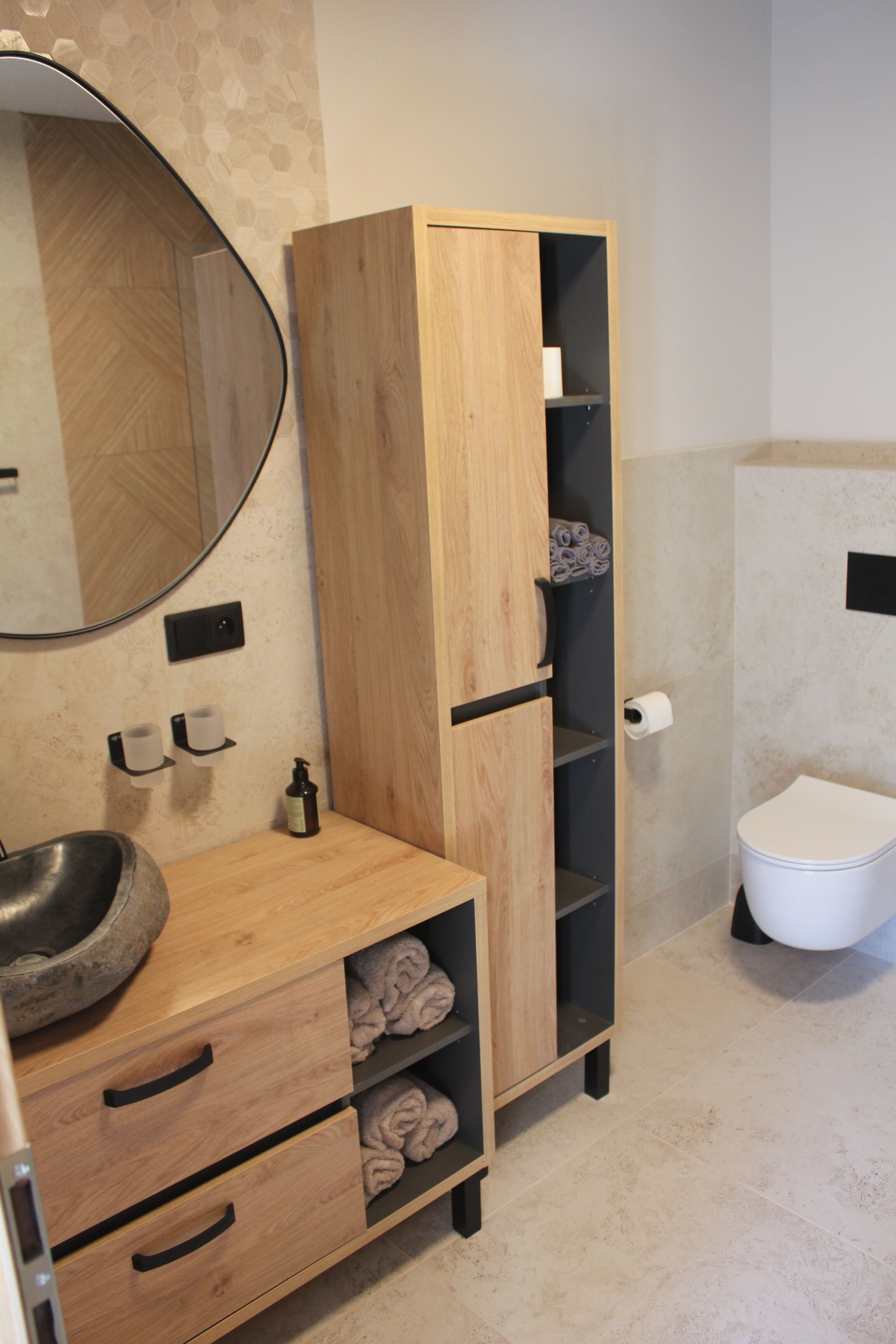 Une salle de bain avec un meuble-lavabo en bois, un miroir rond, des toilettes et un meuble avec des étagères.
