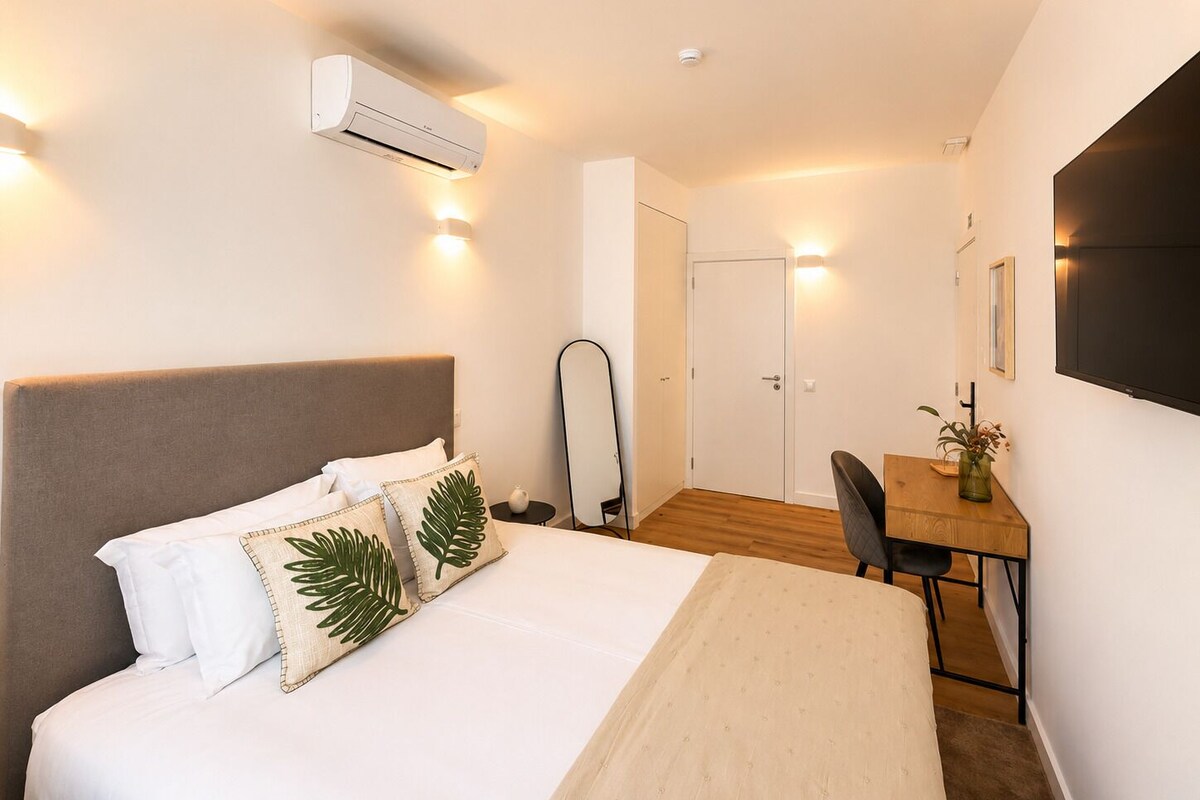 Habitación de hotel con cama, mesita de noche, silla, escritorio, aire acondicionado y una puerta.