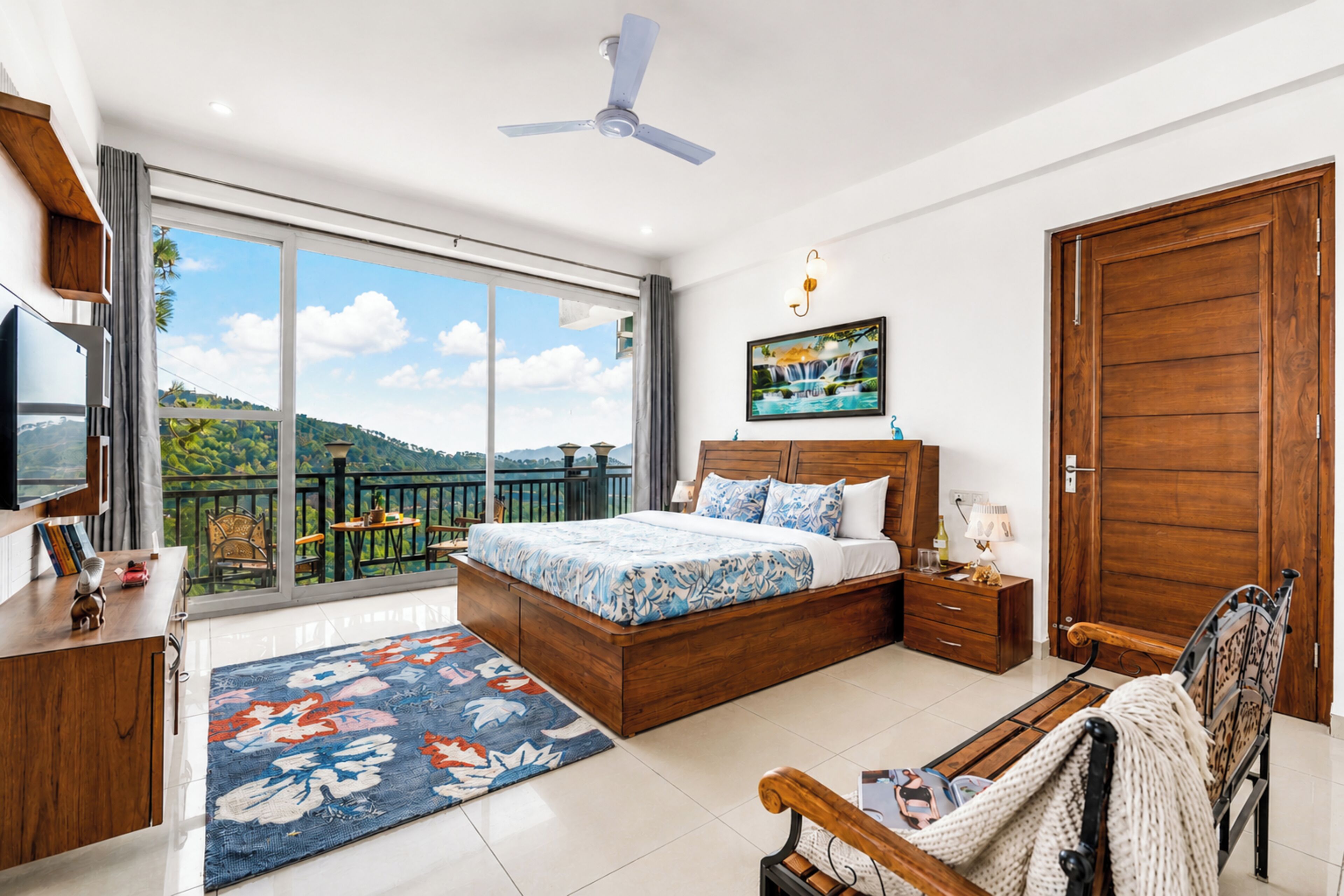 A bedroom with a large bed, a wooden wardrobe, a desk, a chair, and a balcony with a view of the hills.