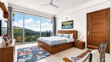 A bedroom with a large bed, a wooden wardrobe, a desk, a chair, and a balcony with a view of the hills.