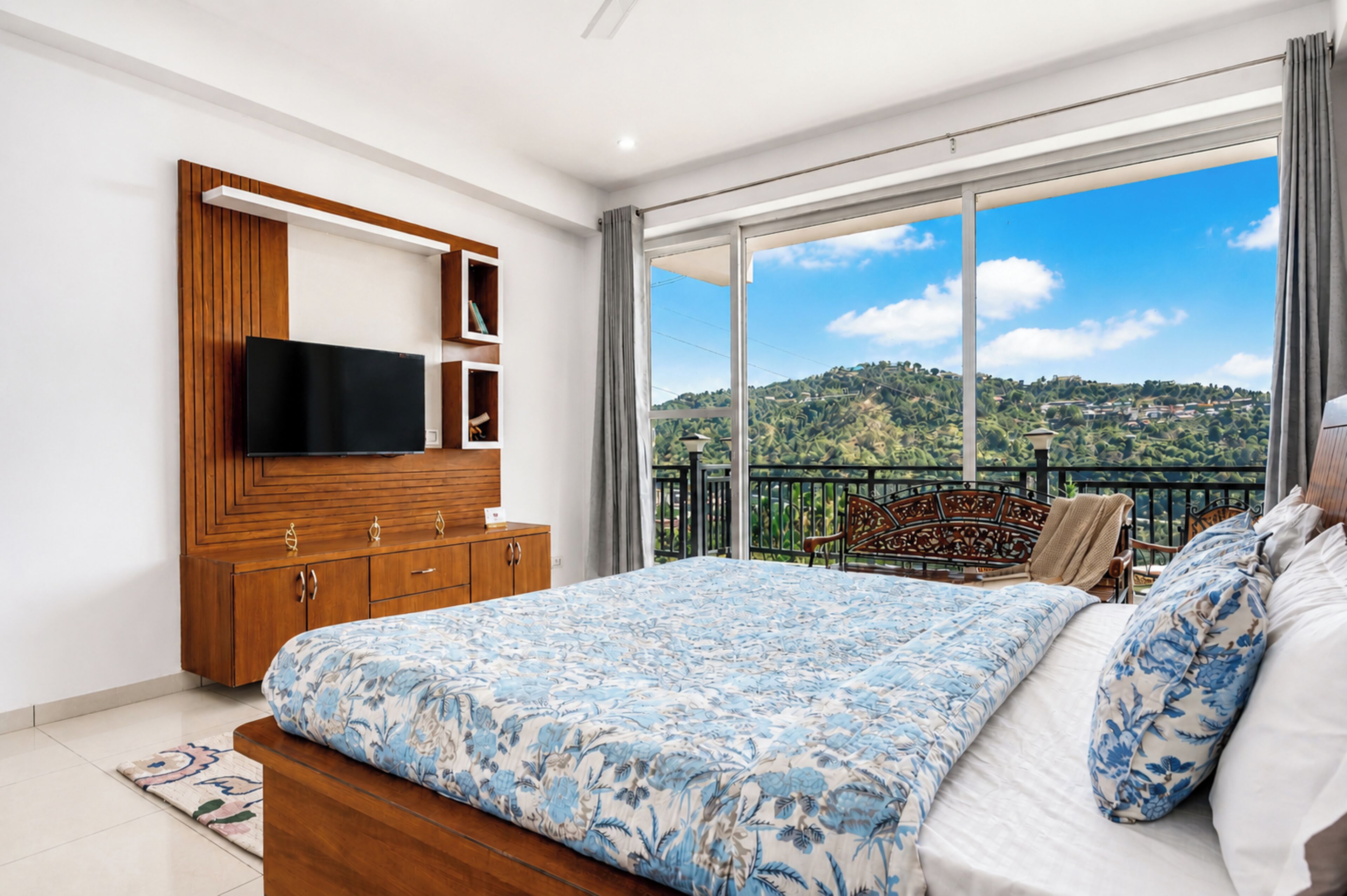 A bedroom with a large bed, a TV mounted on the wall, a wooden cabinet, and a balcony with a view of a green landscape.