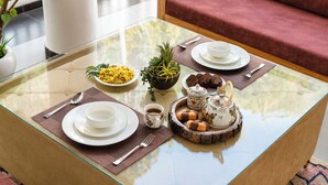 A glass-top dining table with plates, cups, and a tray of pastries, set in a modern room with a red sofa and a potted plant. - Mist & Manor Petfriendly Apartments (Dilman)
